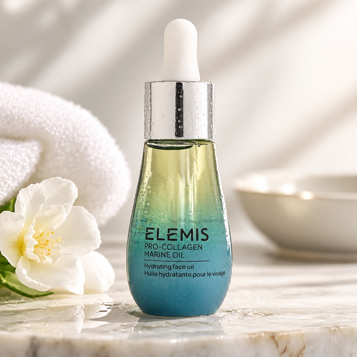 ELEMIS Pro-Collagen Marine Oil in an elegant blue and pale gold glass dropper bottle with a silver cap, beautifully displayed on natural marble beside delicate white flowers and soft towels. Luxurious hydrating facial oil designed to nourish skin, improve the appearance of fine lines and promote a smoother, radiant complexion.