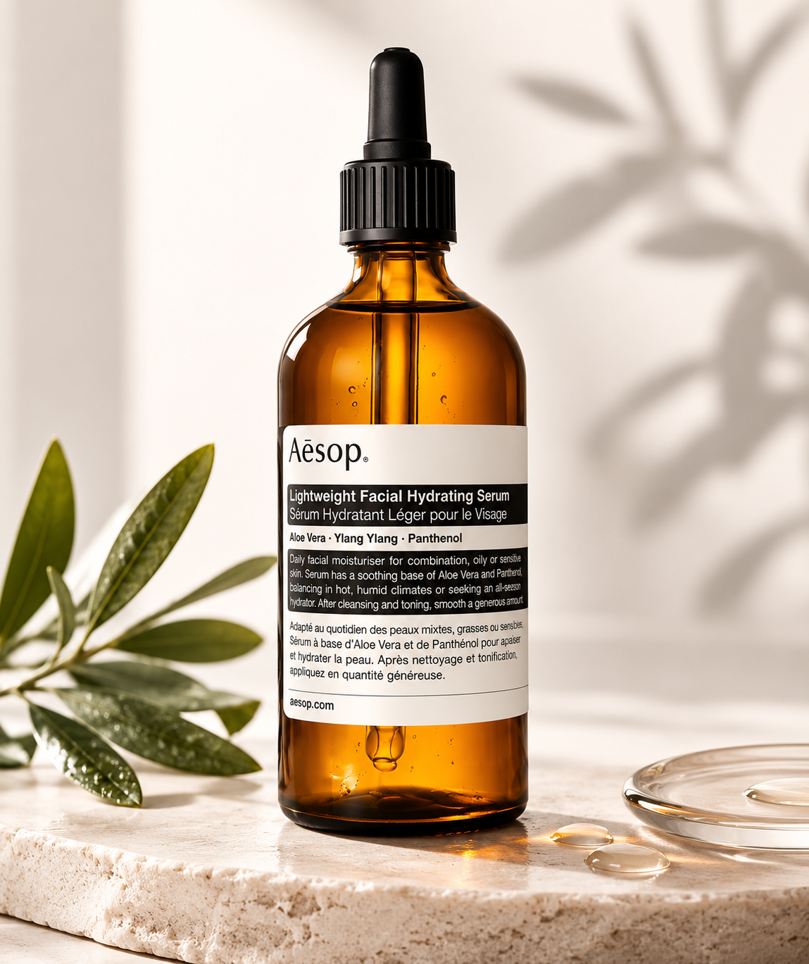 Aesop Lightweight Facial Hydrating Serum displayed on a natural stone countertop in a bright, spa-inspired setting with soft sunlight, olive branches, and glass skincare accessories. The amber glass dropper bottle is styled to emphasize a calming daily skincare ritual, highlighting its lightweight, fast-absorbing formula that hydrates, soothes, and balances combination, oily, and sensitive skin.