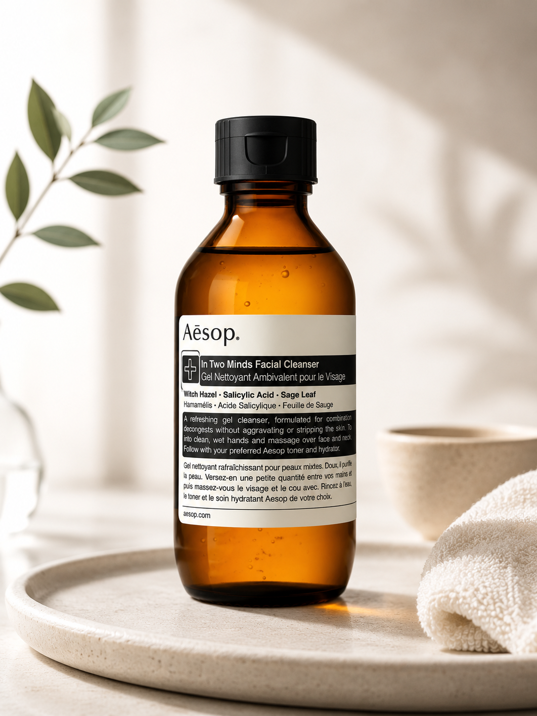 Aesop In Two Minds Facial Cleanser displayed on a minimalist stone tray in a calm, spa-inspired bathroom setting with soft natural light, botanical greenery, a ceramic bowl, and a folded white towel. The warm, clean composition highlights the amber bottle and reflects the cleanser's balancing, purifying, and refreshing benefits for combination skin as part of a modern daily skincare routine.