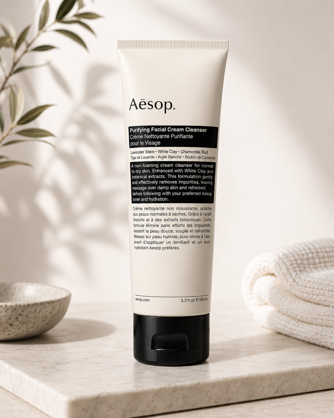 Aesop Purifying Facial Cream Cleanser displayed on a light stone countertop in a minimalist spa-inspired setting with soft natural light, botanical greenery, a ceramic bowl, and neatly folded white towels. The calming composition highlights the gentle cream cleanser as part of a premium skincare routine, emphasizing botanical cleansing, skin comfort, hydration, and Aesop's refined apothecary aesthetic.